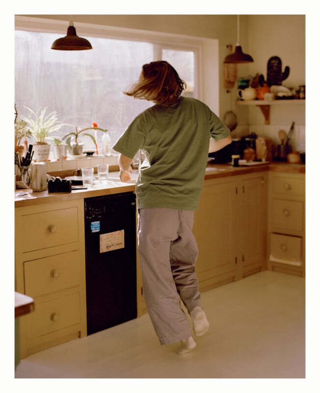 A person with shoulder-length hair, wearing a green t-shirt and loose pants, appears to be dancing or moving energetically in a sunlit kitchen with beige cabinets and plants by the window.