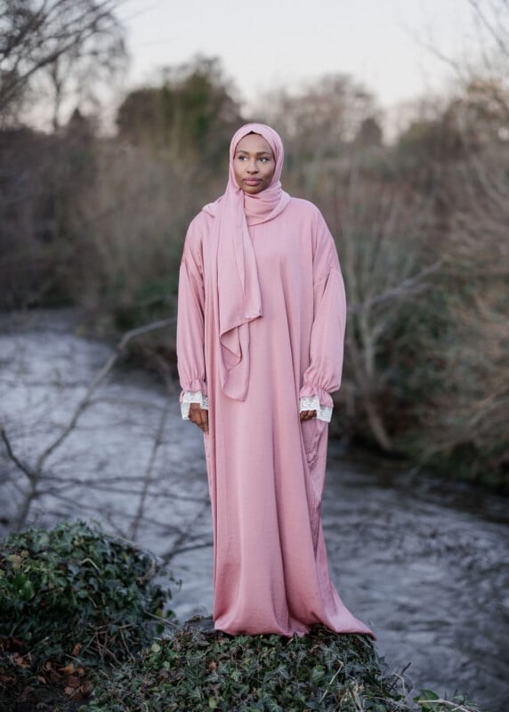 A woman wearing a long, loose-fitting pink dress and matching hijab stands outdoors near a river, surrounded by greenery and bare trees. She looks thoughtfully into the distance.