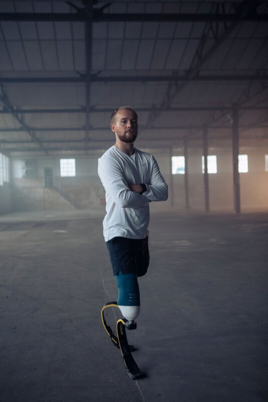 A man with a running blade prosthetic on his right leg stands confidently with arms crossed in a large, empty industrial building. He wears a white long-sleeved shirt and dark shorts.