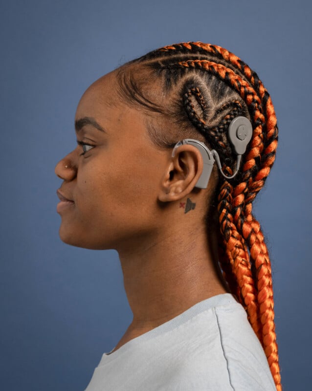 A woman with orange and black braids and a small neck tattoo is shown in profile against a blue background, wearing a cochlear implant device on her ear.