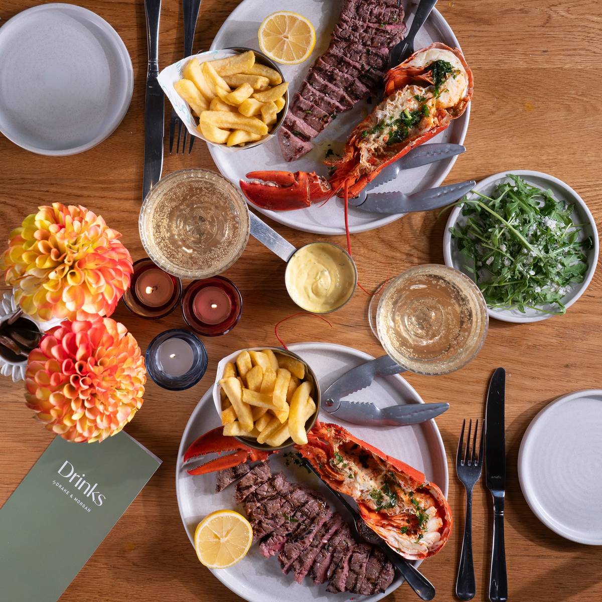 a table set with portions of lobster, steaks, chips, drinks, and side salads