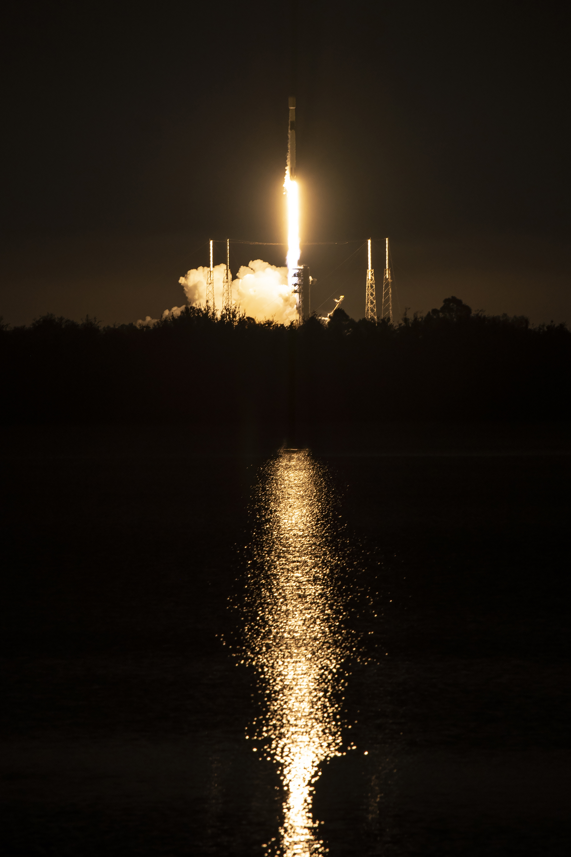 a night launch of an orbital rocket is seen on the horizon and reflected in a water body in the foreground