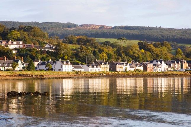 A pretty view of a body of water in the Isle of Arran, with houses sitting on the bank and green trees and hills behind them.