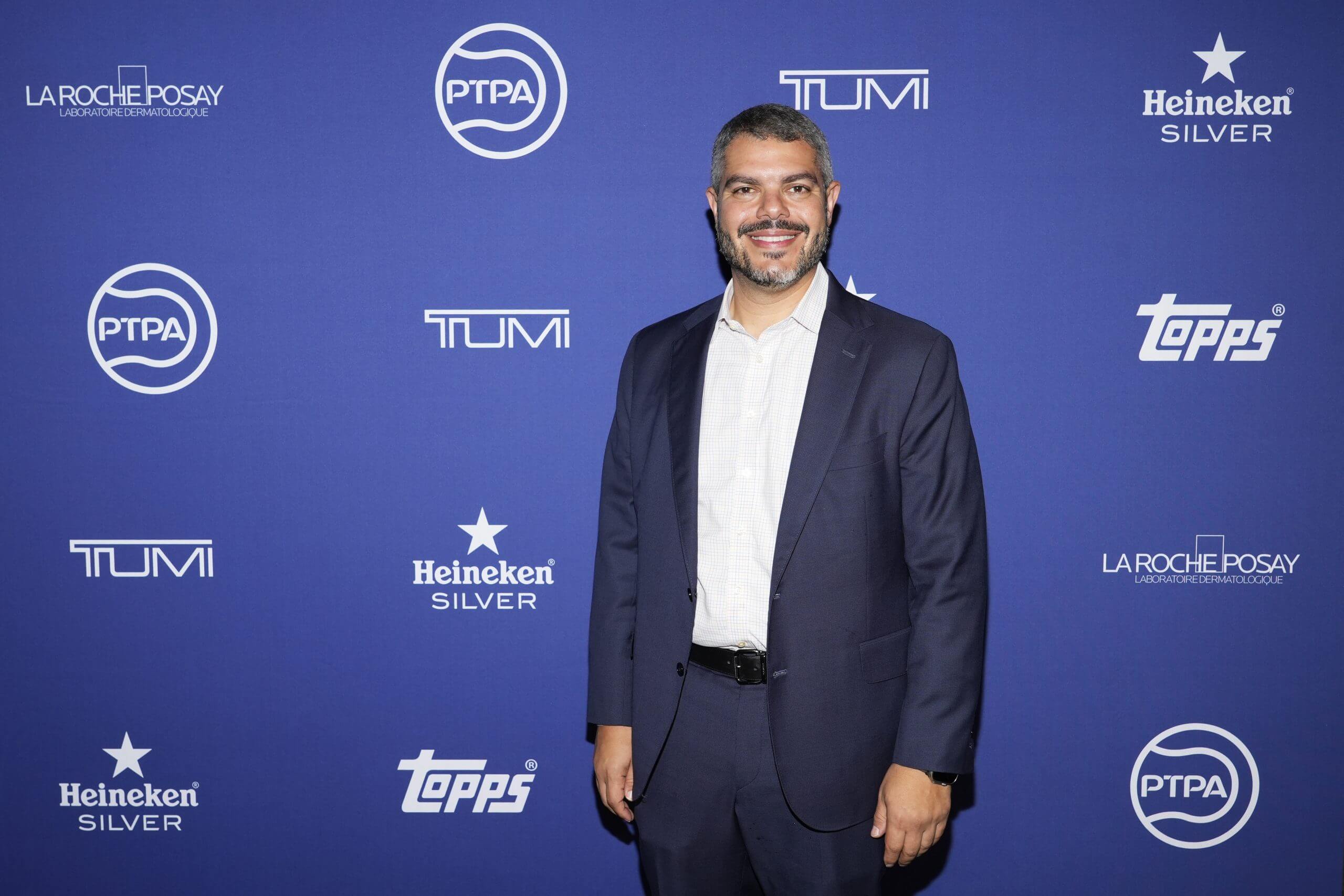 Ahmad Nassar wears a suit in front of a navy backdrop with logos for the Professional Tennis Players' Association, Tumi, Heineken and other sponsors.
