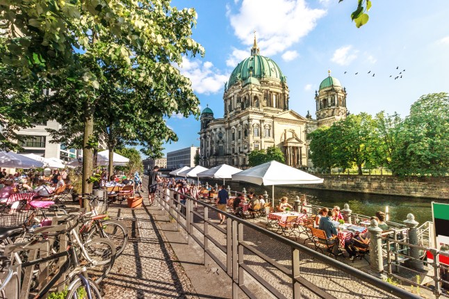 People sitting in outdoor dining areas along the bank of the Spree River in Berlin, Germany, with the Berliner Dom behind it and trees lining a nearby path.