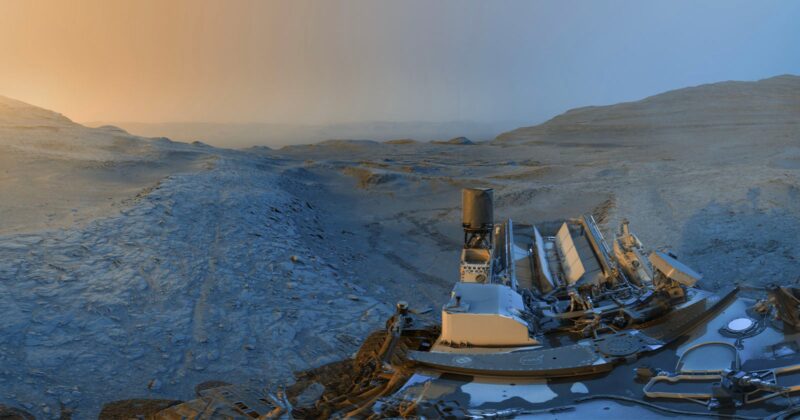 Mars rover sits on rocky terrain under a gradient sky, from orange on the left to blue on the right, with Martian hills and a dusty landscape stretching into the distance.