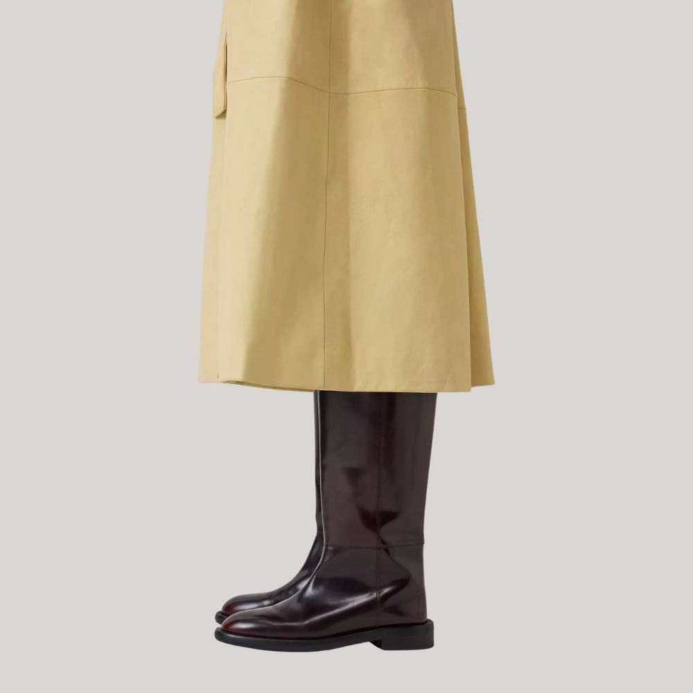 John Lewis x REJINA PYO Knee Length Leather Blend Riding Boots, Burgundy worn with pale yellow trench coat sideways against pale gray background.