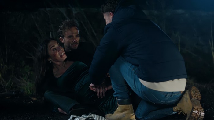 Jacob kneels down to an injured David and Shona in a dark road in a scene from Corriedale