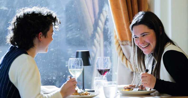 A queer couple has a meal at a fine dining restaurant. One person faces the camera and laughs at the other, who sits on the left away from camera smiling. On the table is food and wine. The person laughing holds a fork in hand and wears a white vest. The other has a black vest.