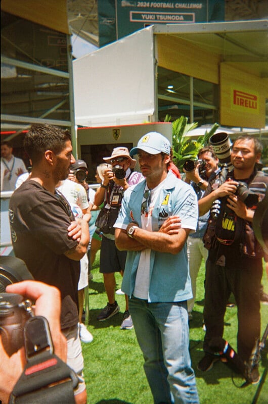Two men stand facing each other with arms crossed, surrounded by photographers and media at an outdoor event. A banner with "2024 Football Challenge" and "Yuki Tsunoda" is visible in the background.