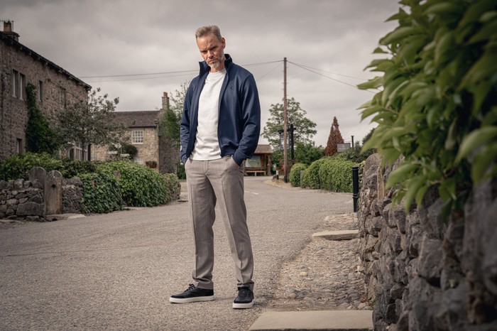 Ray (Joe Absolom) stands with his hands in his pockets in a blue jacket, white t-shirt and grey trousers