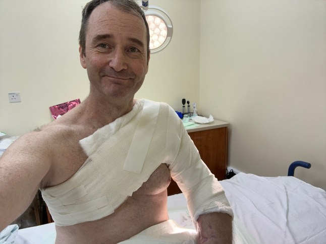 Mark Brooks in a hospital selfie, smiling, with a large bandage over his body and arm, following multiple procedures.