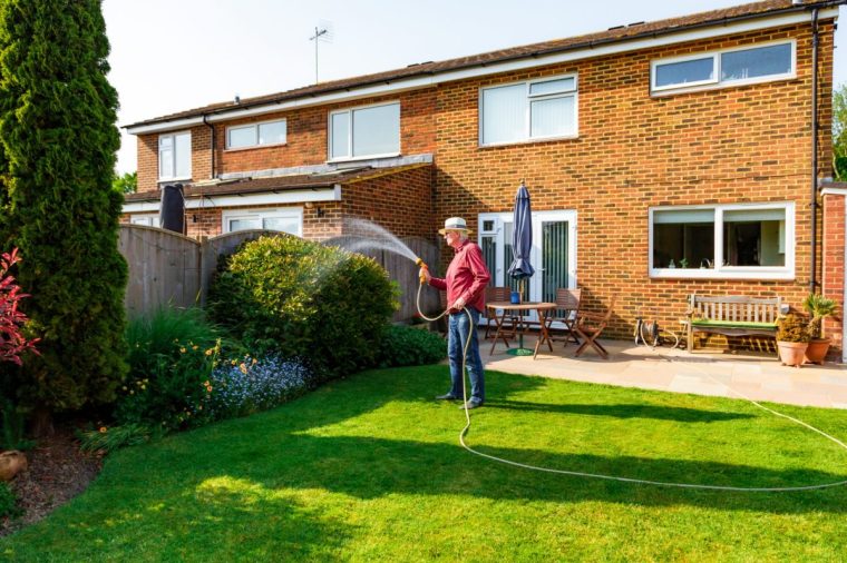 Portrait of a senior man watering his garden using a hose in the summer. It is hot and sunny and the man wears a summer hat to protect himself.