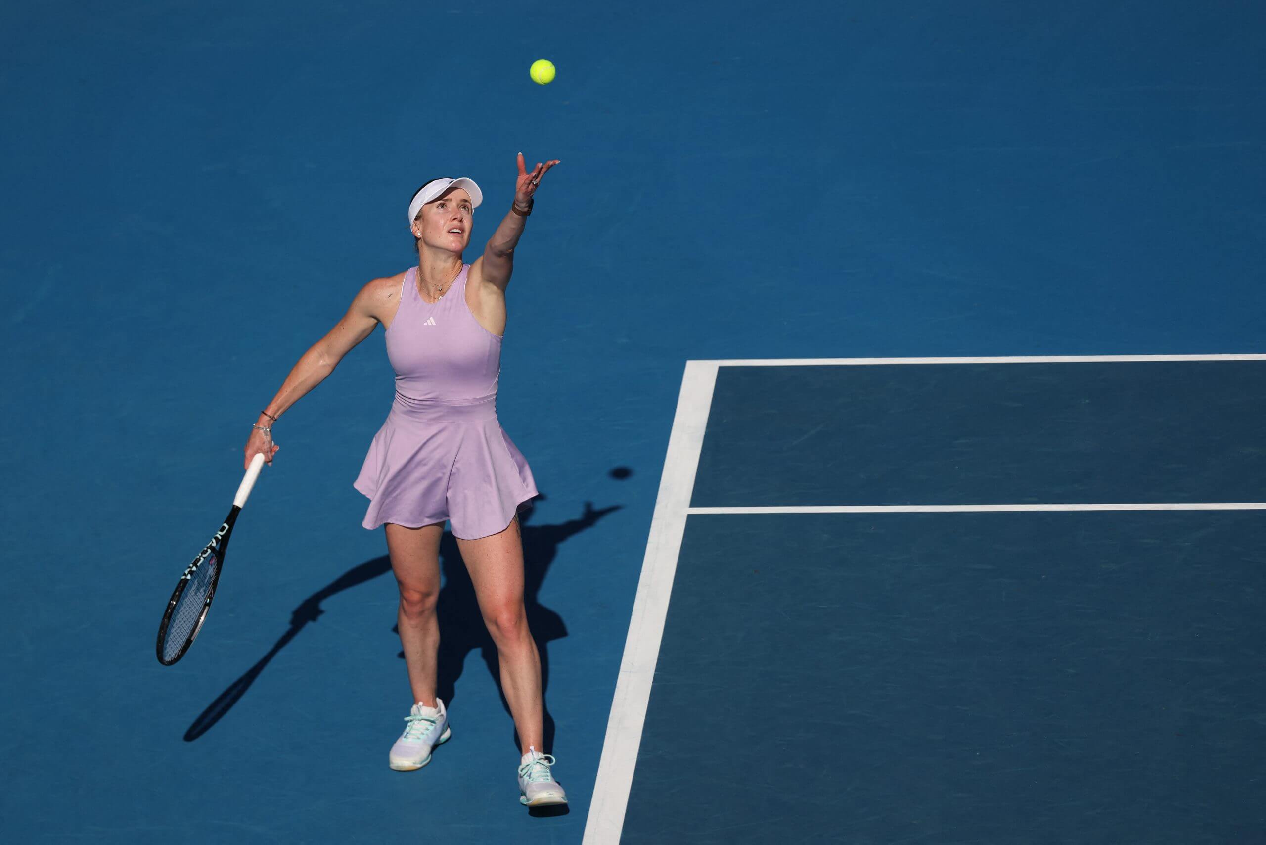 Elina Svitolina tosses the ball in the air to serve on a blue tennis court, wearing a lilac tennis outfit.