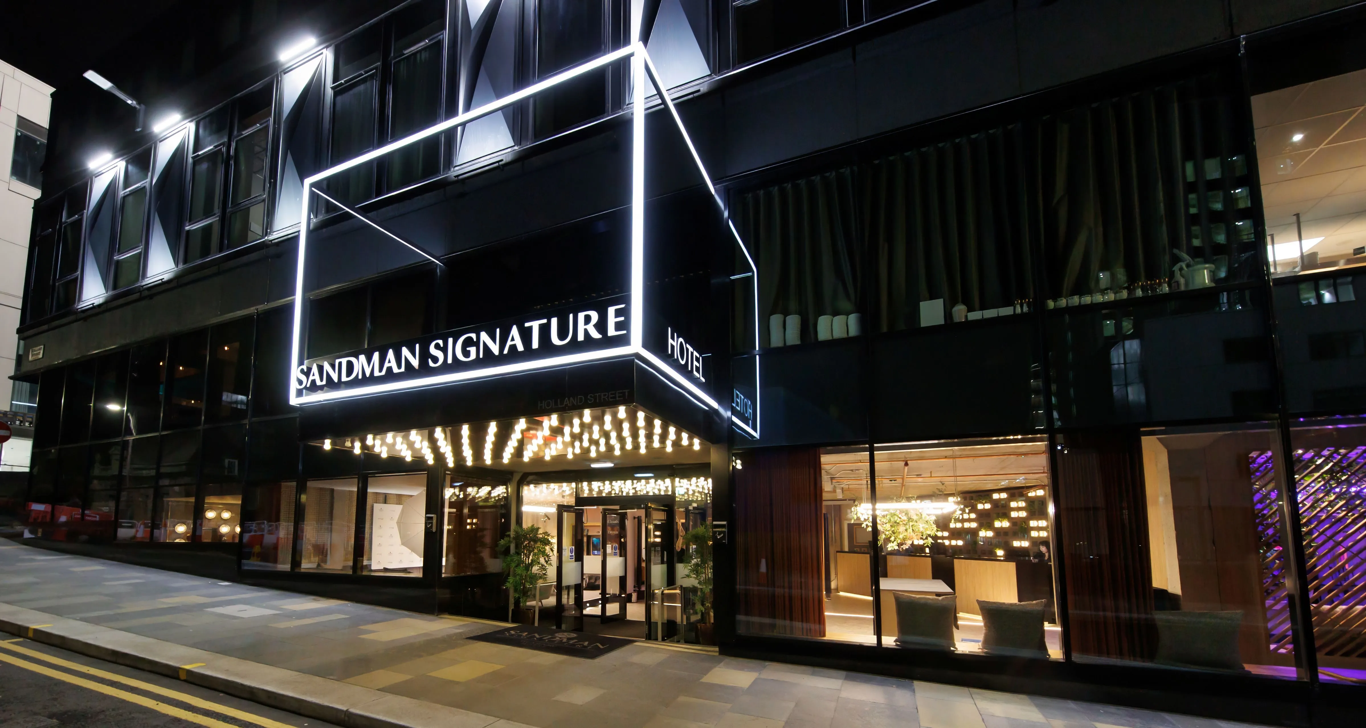 The Sandman Signature Hotel at night, with bright signage and illuminated entrance.