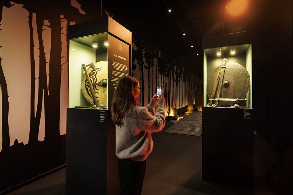 a woman takes a picture with her phone of a display case containing Viking weapons at Vikings: The Immersive Experience