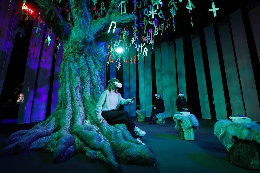 a woman wearing a VR headset sits under a recreation of the mythical Yggdrasil tree at Vikings: The Immersive Experience