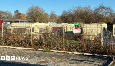 A row of caravans behind a wire fence, with some bushes in the foreground.