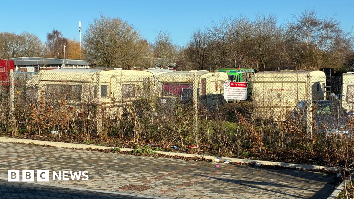 A row of caravans behind a wire fence, with some bushes in the foreground.