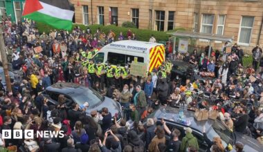 Dozens of people surround an immigration enforcement van on a city street. Several police officers stand in front of the van.