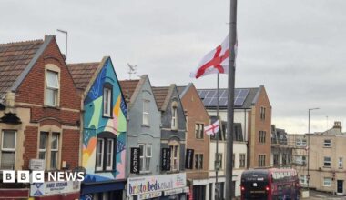 Two St George flags can be seen flying on lampposts. The high street is Duckmoor Road in Bedminster, where there is a row of shops and a bus on the road.