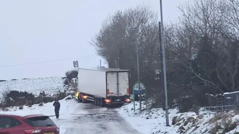 Ruairí McHugh A lorry stuck across a narrow, ice covered rural road in County Tyrone.  The large white vehicle appears to be parked slightly sideways on the road with its lights on.  A man dressed in black is walking on the side of the road and a red car is in the foreground. The ditch is lined with trees and is covered in snow. 