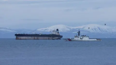 Reuters The tanker is a large ship with rusty marks on its hull. The upper part of its hull is painted blue and it has a white bridge and a single blue funnel. In the foreground is a white US Coast Guard vessel. There are snow-covered hills in the background.