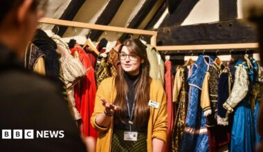 A woman with long brown hair and glasses is wearing a mustard yellow cardigan, black top and yellow and black skirt. she is standing i front of two racks of Tudor costumes