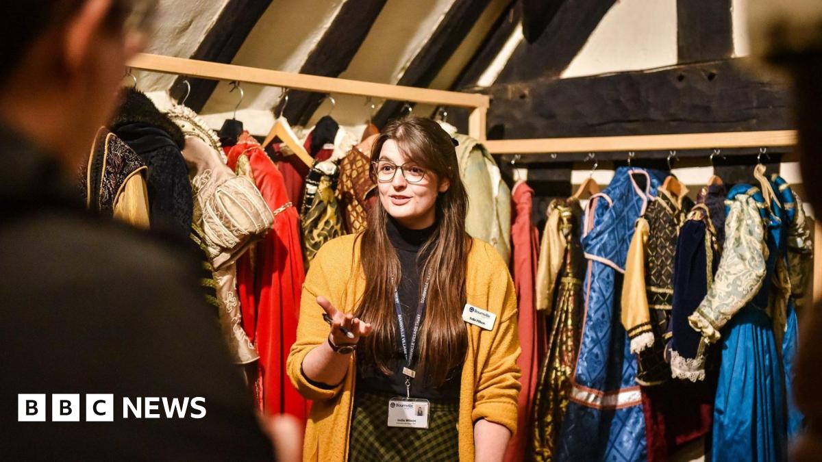 A woman with long brown hair and glasses is wearing a mustard yellow cardigan, black top and yellow and black skirt. she is standing i front of two racks of Tudor costumes