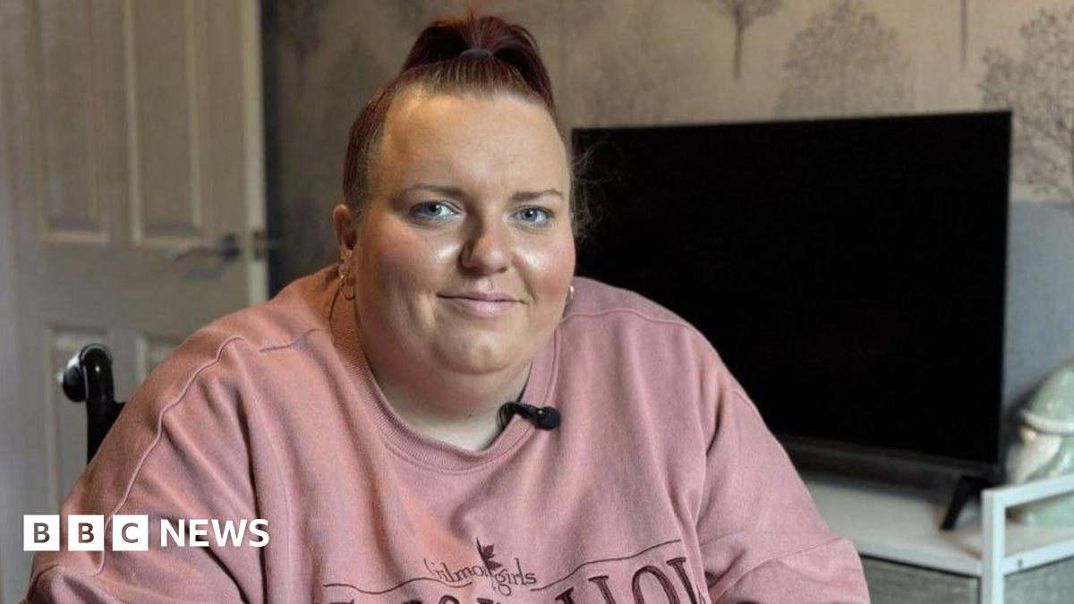Emily Salter, who is wearing a pink jumper, is in her wheelchair in her living room. There is a television on a cabinet and patterned wallpaper in the background. She is smiling at the camera.