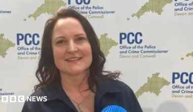 Alison Hernandez wearing a gold necklace and a navy blue top with the top of a Conservative rosette visible and standing in front of a board with the logo for PCC - Office of the Police and Crime Commissioner, Devon and Cornwall.