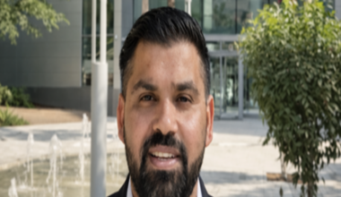 Mann is stood outside a building with a water fountain and small tree behind him. He has a navy suit on and a white shirt. He has dark hair and a beard.