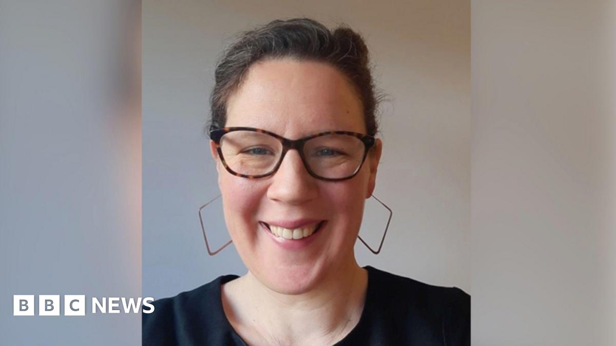 Lucy is looking at the camera and can be seen from the shoulders up. She's wearing large square-hooped earrings and smiling, with her hair back.