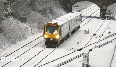A wide shot of a yellow train travelling through snow. The track is covered with snow, and the train is covered with snow.