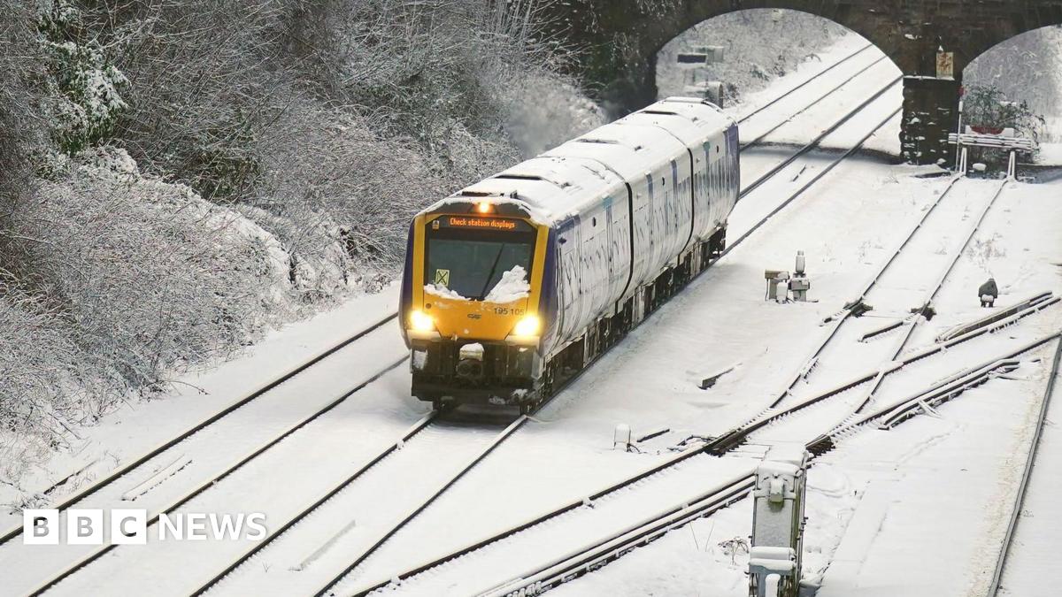 A wide shot of a yellow train travelling through snow. The track is covered with snow, and the train is covered with snow.
