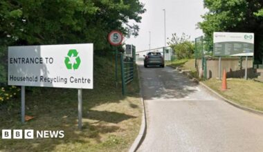 The entrance to Bidston Household Waste Recycling Centre with a large hite sign that says entrance in black writing. A car is travelling along the access road. There is a red circle road sign with 5 written on in black.