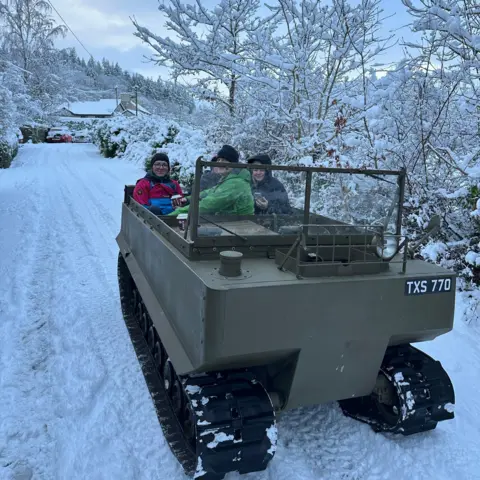 Dawn Stanworth / Anglesey Island A khaki-coloured open-top vehicle, with four people on board, travels along a snowy road. 