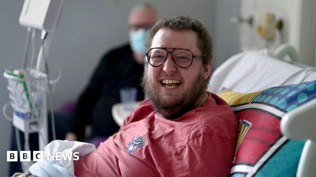 Millie Bobby Brown and David Harbour in character as Eleven and Jim Hopper. Eleven wears red shorts over grey leggings and a grey jumper. Jim Hopper wears a black leather jacket, a black beanie hat and dark trousers.They are speaking to director Frank Darabont on the set of Stranger Things. There is a vintage advertisement posted in the background with debrief on the ground.