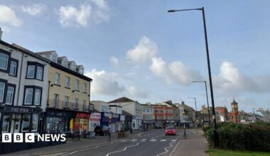 Street view picture of Morecambe. A row of shops are on the left of the road, whilst a grassy verge is on the right. A car drives past the camera and it's a blue sky.