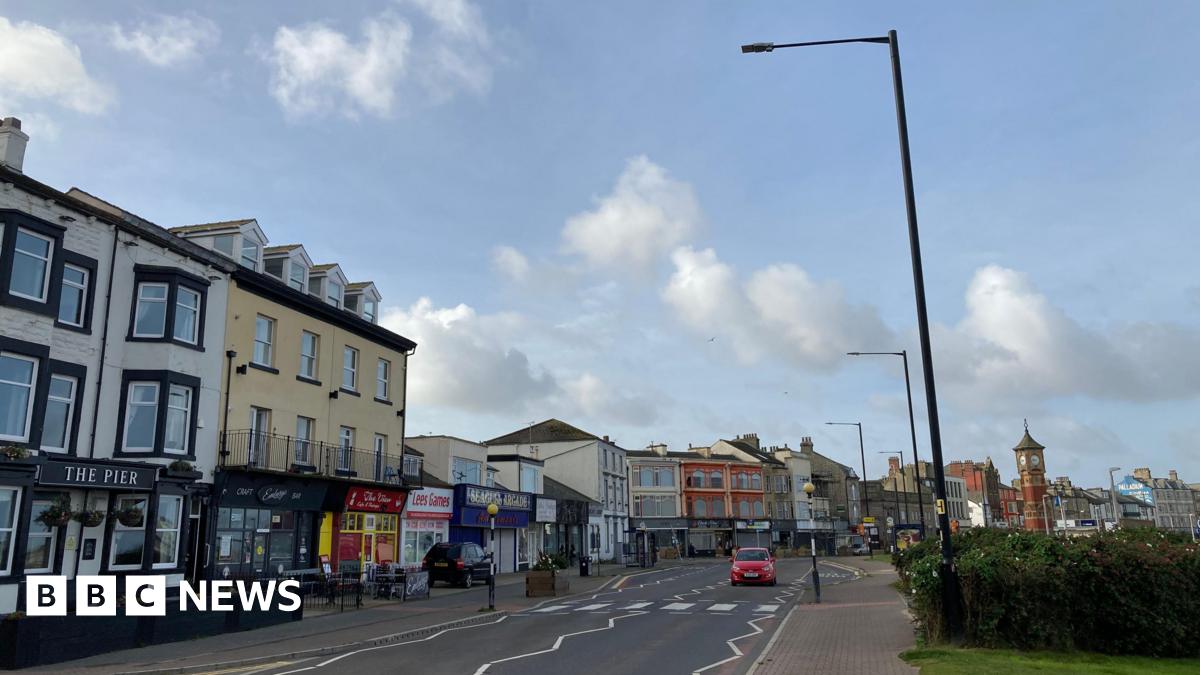Street view picture of Morecambe. A row of shops are on the left of the road, whilst a grassy verge is on the right. A car drives past the camera and it's a blue sky.