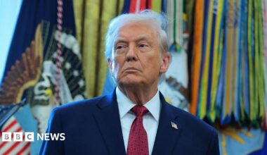Donald Trump stands in the Oval Office, with flags in the background.