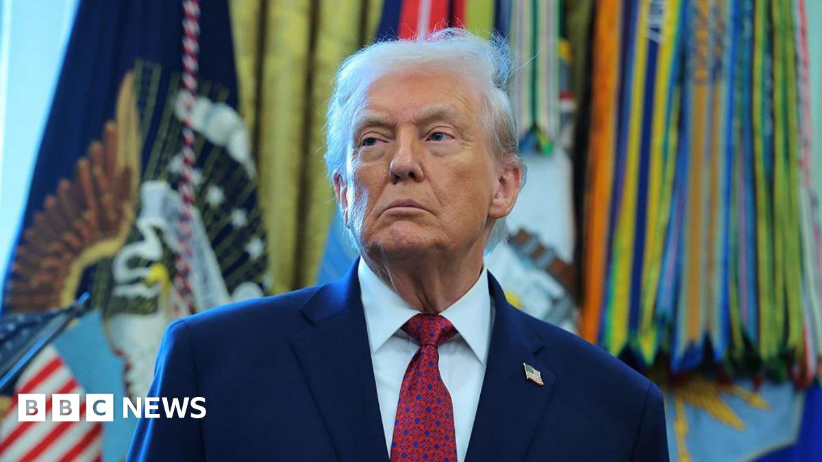 Donald Trump stands in the Oval Office, with flags in the background.