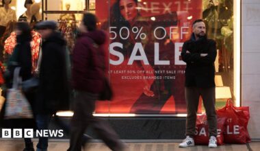 Shoppers walking past a Next store with a red sign in the window saying "50% off sale"