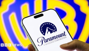 A hand holds a mobile phone, displaying the Paramount logo of a stylised mountain with a halo of stars. In the background is a bright blue and yellow Warner Bros shield logo.