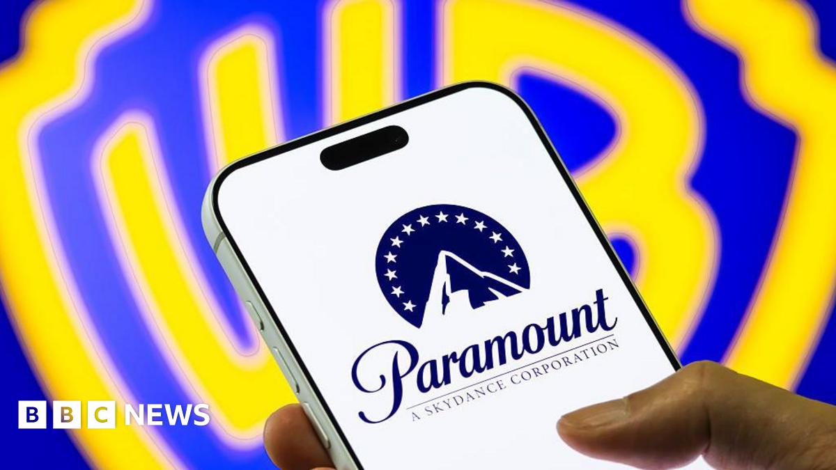 A hand holds a mobile phone, displaying the Paramount logo of a stylised mountain with a halo of stars. In the background is a bright blue and yellow Warner Bros shield logo.