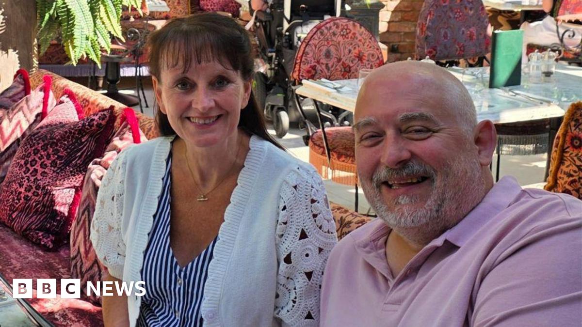 Sue and Dave Cook are sitting in an outdoor area at a restaurant. Sue has long brown hair and is wearing a blue-and-white striped top and a white cardigan. Dave is bald and has a grey beard and is wearing a pink polo shirt. They are both smiling for the camera.