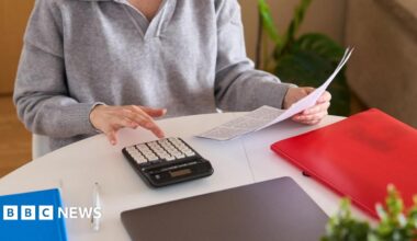 Shona Robison, who has blonde hair tied back, speaks in the Scottish Parliament chamber with her right hand held out in front of her, holding a pen. She is wearing a pink suit jacket and a black and white scarf.