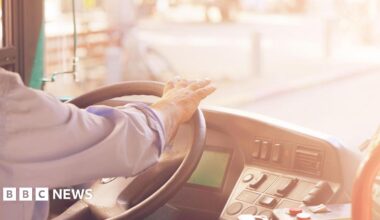 A person wearing a blue shirt has their hands on the driving wheel of a bus. The dashboard of the bus can be seen and a blurred but sunny road out of the front window.