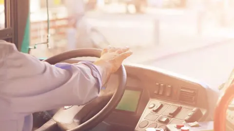 Getty Images A person wearing a blue shirt has their hands on the driving wheel of a bus. The dashboard of the bus can be seen and a blurred but sunny road out of the front window.