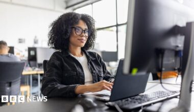 Businesswoman wearing black glasses with dark hair, sitting working on computer at her desk in office